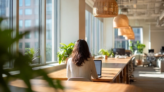 Regus Alternative- What You Actually Need vs What Youre Overpaying For | Alliance Virtual Offices Woman working on a laptop at a shared wood desk in a bright coworking space with large windows and hanging wicker pendant lights.