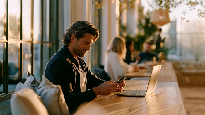Best Virtual Phone Number for Small Business- Top Options Compared | Alliance Virtual Offices Man using his phone next to an open laptop at a sunlit café counter with people in the background