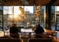 Two people working on laptops at shared tables in a sunlit coworking space