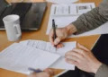 Person signing a contract with a black pen at a wooden desk, with paperwork, a laptop and a white coffee mug nearby