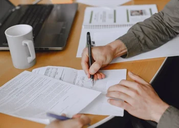 Virtual Address for LLC Registration- Whats Allowed | Alliance Virtual Offices Person signing a contract with a black pen at a wooden desk, with paperwork, a laptop and a white coffee mug nearby