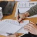 Person signing a contract with a black pen at a wooden desk, with paperwork, a laptop and a white coffee mug nearby