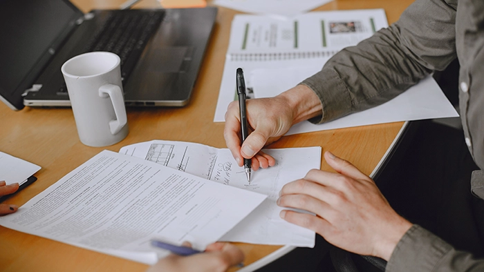 Virtual Address for LLC Registration- Whats Allowed | Alliance Virtual Offices Person signing a contract with a black pen at a wooden desk, with paperwork, a laptop and a white coffee mug nearby