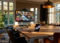Modern meeting room with a large wooden conference table, laptops, and a wall-mounted screen showing a video call grid, lit by natural light from tall windows