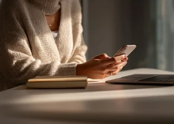 Mobile professional checking her smartphone at a desk with a laptop and notebook, illustrating how virtual receptionist services support US professionals on the go.