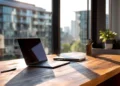 Laptop on a wooden desk by large windows with urban city building view, representing upgrading from a home address to a professional virtual office