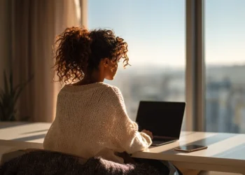 Woman working on laptop at home office desk with city view window, using virtual phone system for small business