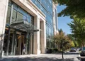 Professional entering the glass-canopied entrance of a modern office building with a stone and glass facade, lined by mature trees and a landscaped sidewalk on a sunny day