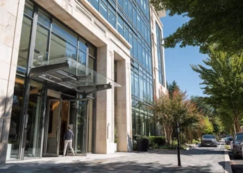 Professional entering the glass-canopied entrance of a modern office building with a stone and glass facade, lined by mature trees and a landscaped sidewalk on a sunny day