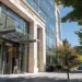 How to Choose an Enterprise Virtual Office Solution in 2026 | Alliance Virtual Offices Professional entering the glass-canopied entrance of a modern office building with a stone and glass facade, lined by mature trees and a landscaped sidewalk on a sunny day