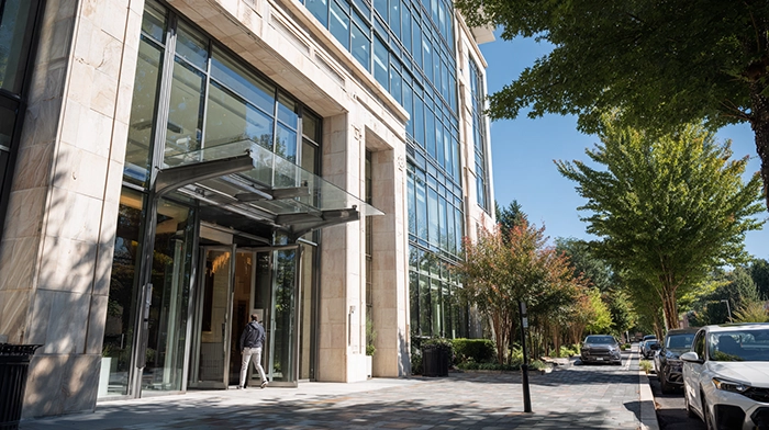 How to Choose an Enterprise Virtual Office Solution in 2026 | Alliance Virtual Offices Professional entering the glass-canopied entrance of a modern office building with a stone and glass facade, lined by mature trees and a landscaped sidewalk on a sunny day
