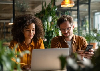 Two remote professionals collaborating at a laptop in a modern coworking space filled with green plants, representing virtual office solutions for distributed teams.