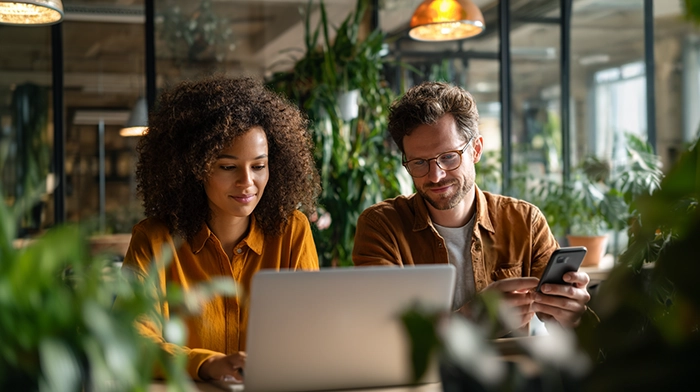 Virtual Office Solutions for Remote Teams- Your 2026 Guide to Working Professionally from Anywhere | Alliance Virtual Offices Two remote professionals collaborating at a laptop in a modern coworking space filled with green plants, representing virtual office solutions for distributed teams.