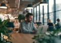 Man working on a laptop at a shared table in a modern coworking space with industrial lighting, large windows, and other professionals in the background