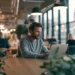 Man working on a laptop at a shared table in a modern coworking space with industrial lighting, large windows, and other professionals in the background