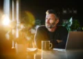 Small business owner sitting at a desk with a laptop and coffee mug, looking confident while considering AI answering service options for his company