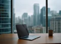 Laptop and coffee mug on a wooden desk in a high-rise office with a panoramic city skyline view through floor-to-ceiling windows on a cloudy day