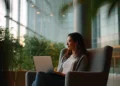 Woman working on a laptop in a modern office lounge with floor-to-ceiling windows and indoor plants