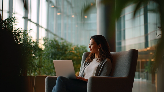 Virtual Office Solutions for Growing Businesses | Alliance Virtual Offices Woman working on a laptop in a modern office lounge with floor-to-ceiling windows and indoor plants