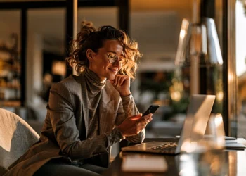 Best Business Phone Number Apps for Small Business in 2026 | Alliance Virtual Offices Woman in glasses and a blazer smiling as she looks at her smartphone while working at a laptop in a warmly lit home office at sunset