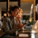 Best Business Phone Number Apps for Small Business in 2026 | Alliance Virtual Offices Woman in glasses and a blazer smiling as she looks at her smartphone while working at a laptop in a warmly lit home office at sunset