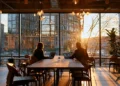 Two people working on laptops at a shared coworking table by large windows