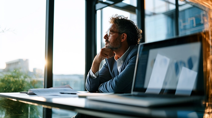How to Start a Real Estate Investment Business in 2026 | Alliance Virtual Offices Man with glasses seated at a desk beside a floor-to-ceiling window, laptop and papers in front of him, looking out at the city
