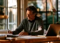 Man writing on paperwork beside a laptop at a home office desk by a sunlit window