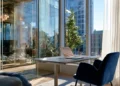 Sunlit private office with a laptop on a height-adjustable desk, navy velvet chairs, and floor-to-ceiling windows overlooking a city street