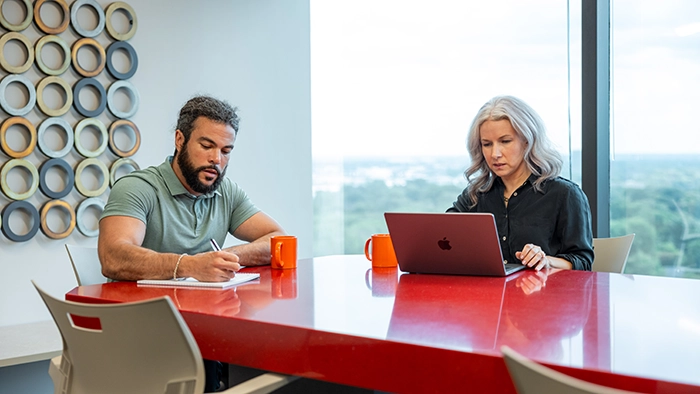 LLC Annual Requirements- A Yearly Compliance Checklist to Stay in Good Standing | Alliance Virtual Offices Two professionals working at a red conference table in a modern Alliance Virtual Offices meeting room with floor-to-ceiling windows