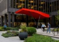 Pedestrians walking past the landscaped plaza entrance of a modern office tower with a bright red canopy