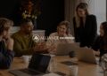 Five colleagues collaborating around a conference table with laptops and coffee mugs in a modern office, with the Alliance Enterprise logo centered