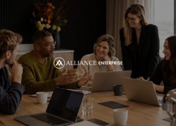 Five colleagues collaborating around a conference table with laptops and coffee mugs in a modern office, with the Alliance Enterprise logo centered