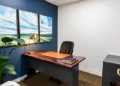 Private day office with a wood desk, black leather chair, brass desk lamp, and coastal artwork on a navy accent wall