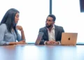 Two business professionals talking at a conference table with laptops in a bright meeting room